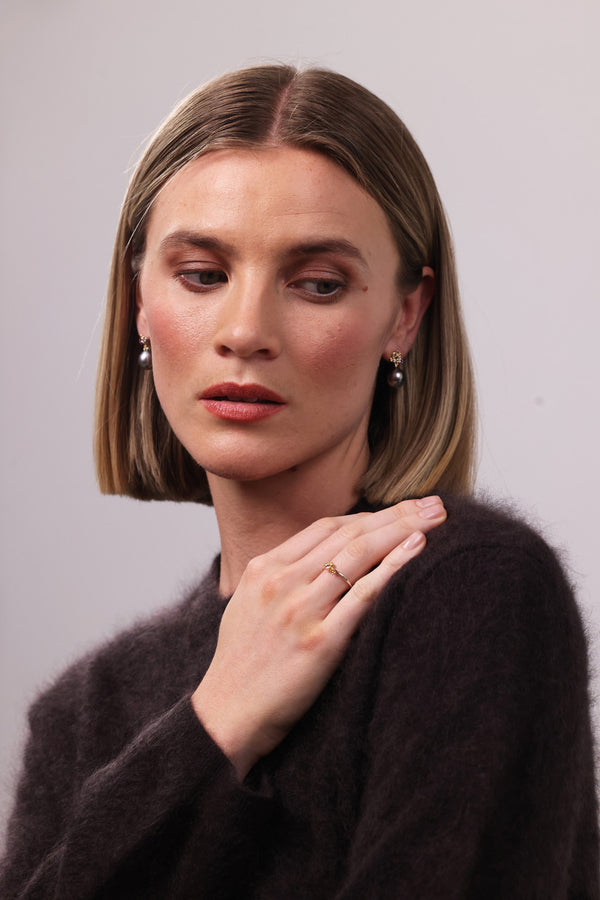 Woman with short blonde hair wearing a dark brown sweater against a plain background wearing pearl earrings